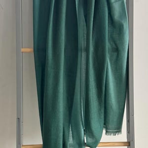 May include: A dark green, soft, and lightweight scarf hanging on a wooden rack. The scarf has a fringed edge and a small tag with the word "Cashmere" on it.