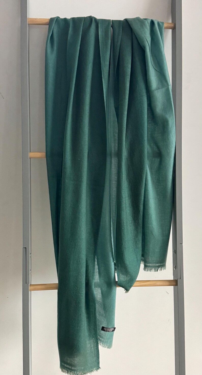 May include: A dark green, soft, and lightweight scarf hanging on a wooden rack. The scarf has a fringed edge and a small tag with the word "Cashmere" on it.