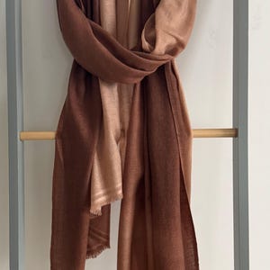 以下が含まれることがあります： フリンジの縁取りが施された、茶色と桃色のグラデーションのストール。ストールは木の棒にかけられています。