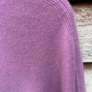 May include: A mauve-colored knitted sweater featuring a fine, uniform stockinette stitch pattern. The garment displays a rounded neckline and a structured shoulder seam, showcasing the soft, textured surface of the knit fabric in a solid purple hue.