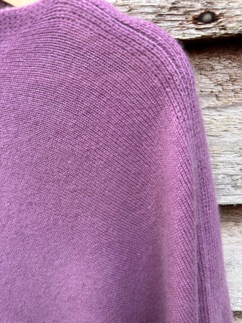 May include: A mauve-colored knitted sweater featuring a fine, uniform stockinette stitch pattern. The garment displays a rounded neckline and a structured shoulder seam, showcasing the soft, textured surface of the knit fabric in a solid purple hue.