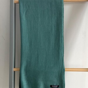 May include: A teal green pashmina scarf with a fringed edge. The scarf is hanging on a wooden rack.