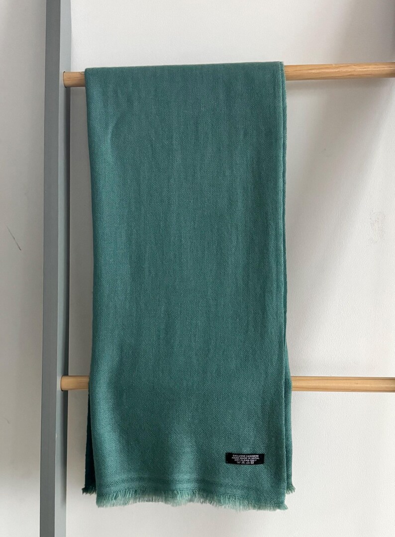May include: A teal green pashmina scarf with a fringed edge. The scarf is hanging on a wooden rack.