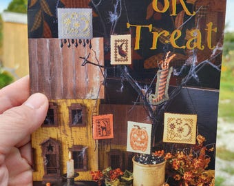 May include: A Halloween-themed book cover with the words "Trix or Treat" in orange. The cover features a miniature house with cross-stitch designs of a pumpkin, a crow, and a witch's stocking. The words "Blackbird Designs" are at the bottom.
