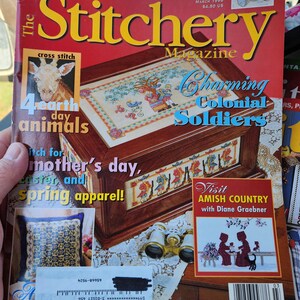 May include: The Stitchery Magazine, March 1998 issue, featuring cross-stitch patterns. The cover displays a wooden box with a cross-stitched design, the text "Charming Colonial Soldiers", and other needlepoint projects. Includes "Eugenie" in cross stitch & needlepoint.