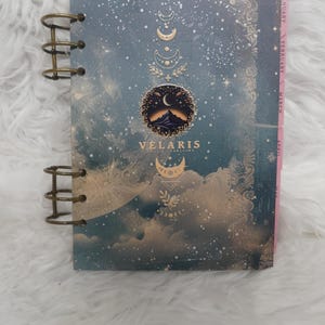 May include: A spiral-bound planner with a dark blue cover featuring gold celestial moon phases, stars, and clouds. The center displays a circular mountain graphic above the text VELARIS. Pink monthly tabs for January through May are visible on the side.