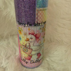 May include: A glittery tumbler with a patchwork design. The tumbler features a purple glitter section, a blue glitter section, and a section with a cartoon duck wearing a pink bow and boots, carrying a bag. Text on the tumbler reads "Sorry I'm late I was Dilly Dallying."