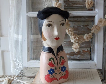 Könnte beinhalten: Ein bemalter Holz-Mannequin-Kopf mit einem schwarzen Hut und einem Blumendesign. Der Mannequin trägt einen schwarzen Hut mit breiter Krempe und ein pinkfarbenes und grünes Blumendesign auf der Brust. Der Mannequin sitzt auf einem Holztisch mit einer weißen Spitzentischdecke.