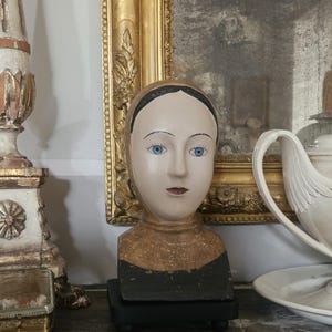 May include: A decorative bust of a person with a pale face, bright blue eyes, and dark painted hair. The figure wears a textured gold-toned head covering and a black base, resting on a small black stand with rounded feet for display purposes.