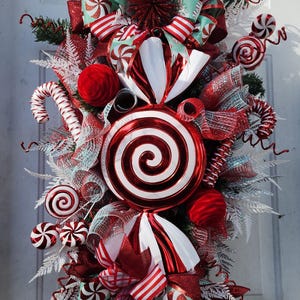 Puede incluir: Una guirnalda de puerta de Navidad con tem&aacute;tica de bast&oacute;n de caramelo rojo y blanco con una gran piruleta roja y blanca en espiral, cinta roja y blanca a rayas y caramelos de menta blanca y roja.