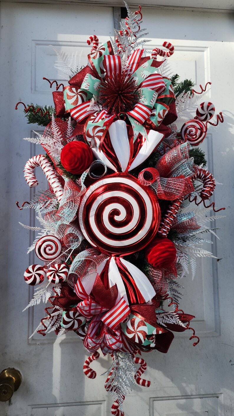 Puede incluir: Una guirnalda de puerta de Navidad con tem&aacute;tica de bast&oacute;n de caramelo rojo y blanco con una gran piruleta roja y blanca en espiral, cinta roja y blanca a rayas y caramelos de menta blanca y roja.