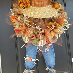 Puede incluir: Una corona de espantap&aacute;jaros caprichosa con un sombrero de paja, cinta a cuadros y piernas de mezclilla. La corona est&aacute; adornada con acentos tem&aacute;ticos de oto&ntilde;o como girasoles, hojas y un peque&ntilde;o cuervo negro. Una encantadora decoraci&oacute;n oto&ntilde;al.