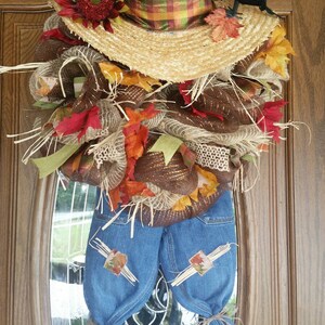 Puede incluir: Una corona de espantap&aacute;jaros hecha a mano con un sombrero de paja, un peto vaquero y decoraciones oto&ntilde;ales. La corona presenta un sombrero de paja con una cinta a cuadros, un cuervo negro y coloridas hojas de oto&ntilde;o. El peto vaquero tiene parches y est&aacute; atado en la parte inferior.
