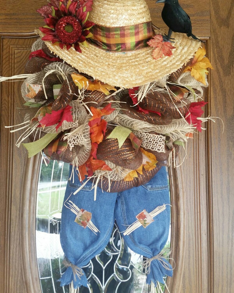 Puede incluir: Una corona de espantap&aacute;jaros hecha a mano con un sombrero de paja, un peto vaquero y decoraciones oto&ntilde;ales. La corona presenta un sombrero de paja con una cinta a cuadros, un cuervo negro y coloridas hojas de oto&ntilde;o. El peto vaquero tiene parches y est&aacute; atado en la parte inferior.