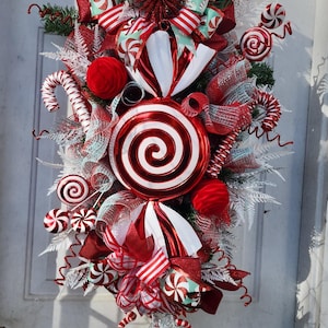 Puede incluir: Una corona de Navidad de bast&oacute;n de caramelo rojo y blanco con una gran piruleta roja y blanca en el centro. La corona est&aacute; decorada con cinta roja y blanca, bastones de caramelo rojo y blanco y purpurina blanca y plateada.