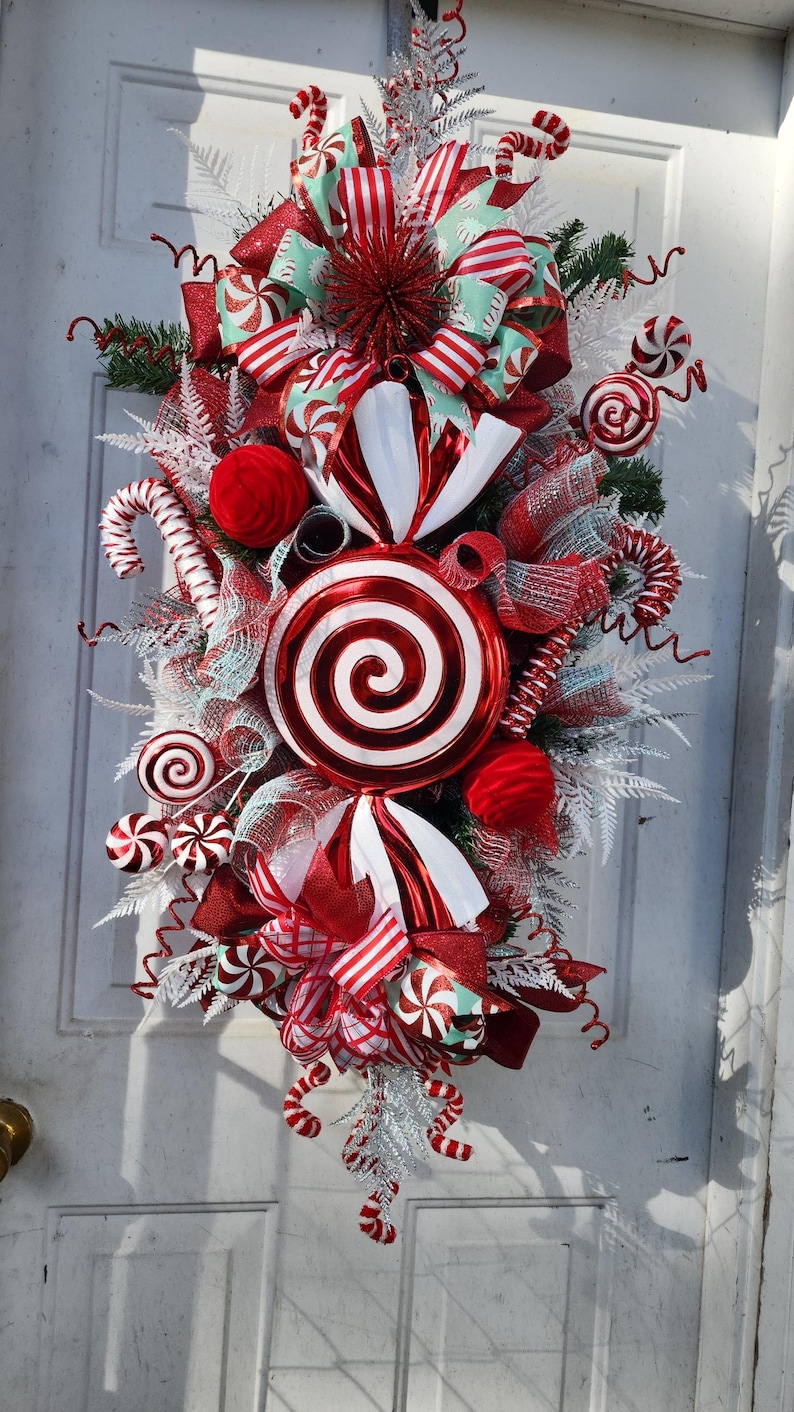 Puede incluir: Una corona de Navidad de bast&oacute;n de caramelo rojo y blanco con una gran piruleta roja y blanca en el centro. La corona est&aacute; decorada con cinta roja y blanca, bastones de caramelo rojo y blanco y purpurina blanca y plateada.