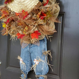 Puede incluir: Una corona de espantap&aacute;jaros con tem&aacute;tica oto&ntilde;al con un sombrero de paja, un cuervo negro y vaqueros de mezclilla. La corona est&aacute; adornada con hojas de oto&ntilde;o, girasoles y malla marr&oacute;n. Los vaqueros tienen detalles de paja y est&aacute;n atados con hilo.