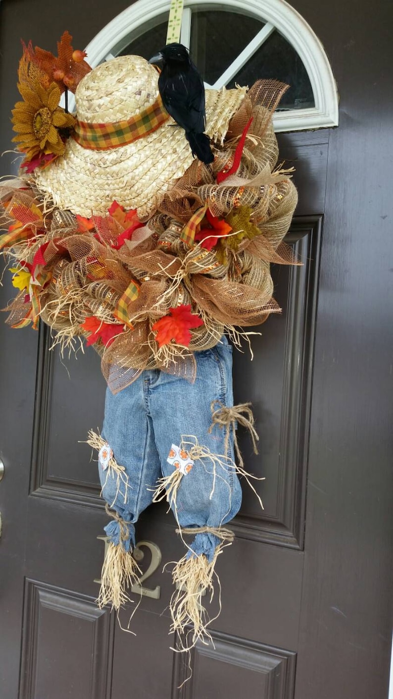 Puede incluir: Una corona de espantap&aacute;jaros con tem&aacute;tica oto&ntilde;al con un sombrero de paja, un cuervo negro y vaqueros de mezclilla. La corona est&aacute; adornada con hojas de oto&ntilde;o, girasoles y malla marr&oacute;n. Los vaqueros tienen detalles de paja y est&aacute;n atados con hilo.