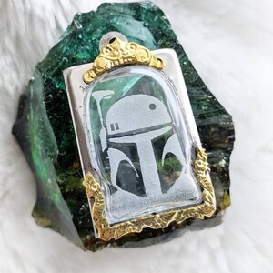 May include: A silver and gold pendant featuring a clear glass cameo with a white silhouette of a Star Wars character's helmet. The pendant is set against a green and gold background.