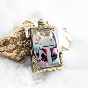 May include: A silver and gold pendant with a clear glass insert featuring a white etched design of a Star Wars character's helmet. The glass insert is filled with colorful beads.