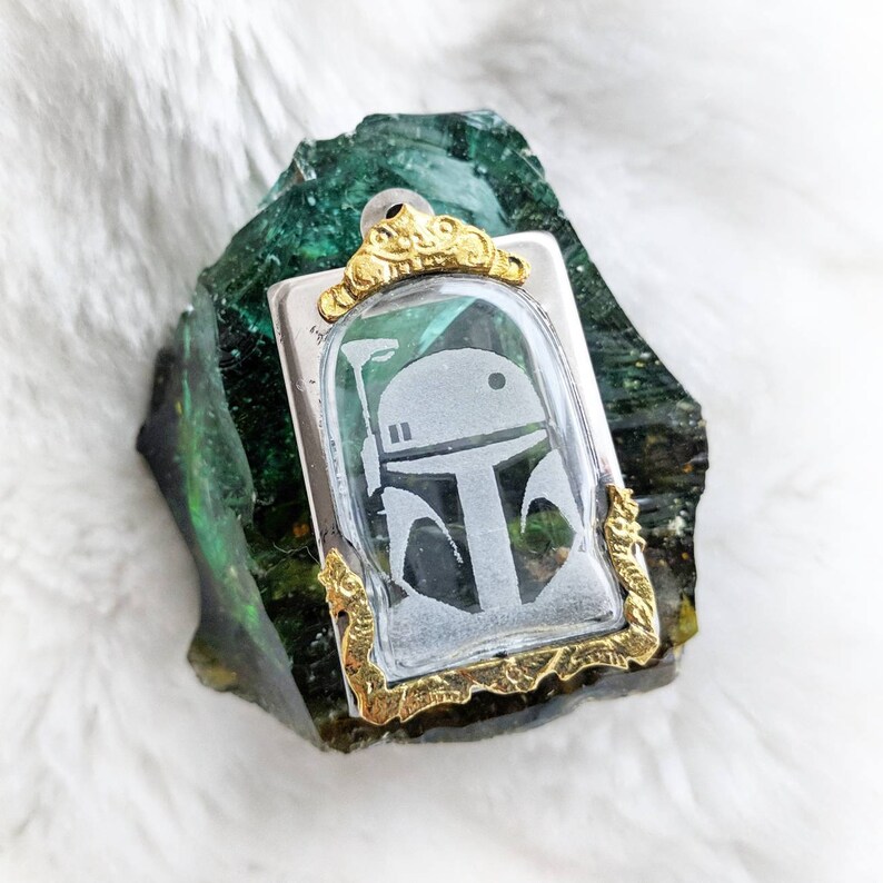 May include: A silver and gold pendant featuring a clear glass cameo with a white silhouette of a Star Wars character's helmet. The pendant is set against a green and gold background.