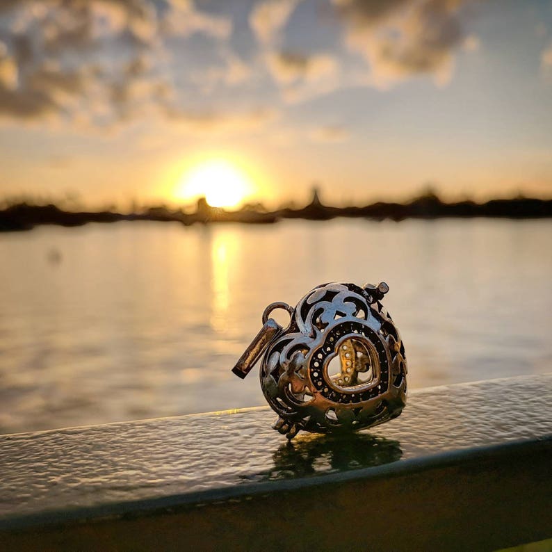 May include: A silver heart-shaped charm with intricate details hangs on a railing against a blurry sunset background.