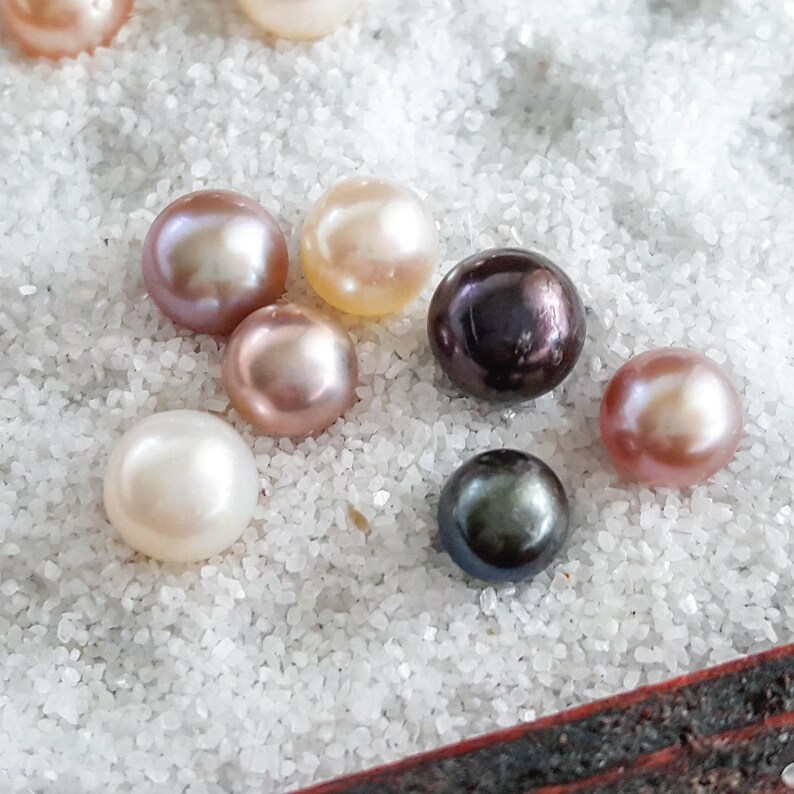 May include: A collection of pearls in various colors, including white, pink, and black, displayed on a white sandy surface. The pearls are round and smooth, with a subtle iridescent sheen.