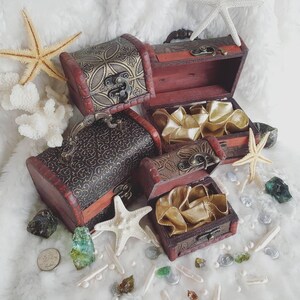 May include: Four ornate wooden treasure chests with intricate designs and brass accents. The chests are open and reveal a golden satin lining. The chests are surrounded by seashells, starfish, and other nautical decor.