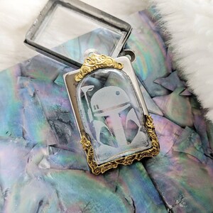 May include: A silver and gold pendant with a clear glass insert featuring an etched image of a Star Wars character's helmet. The pendant is on a white background.