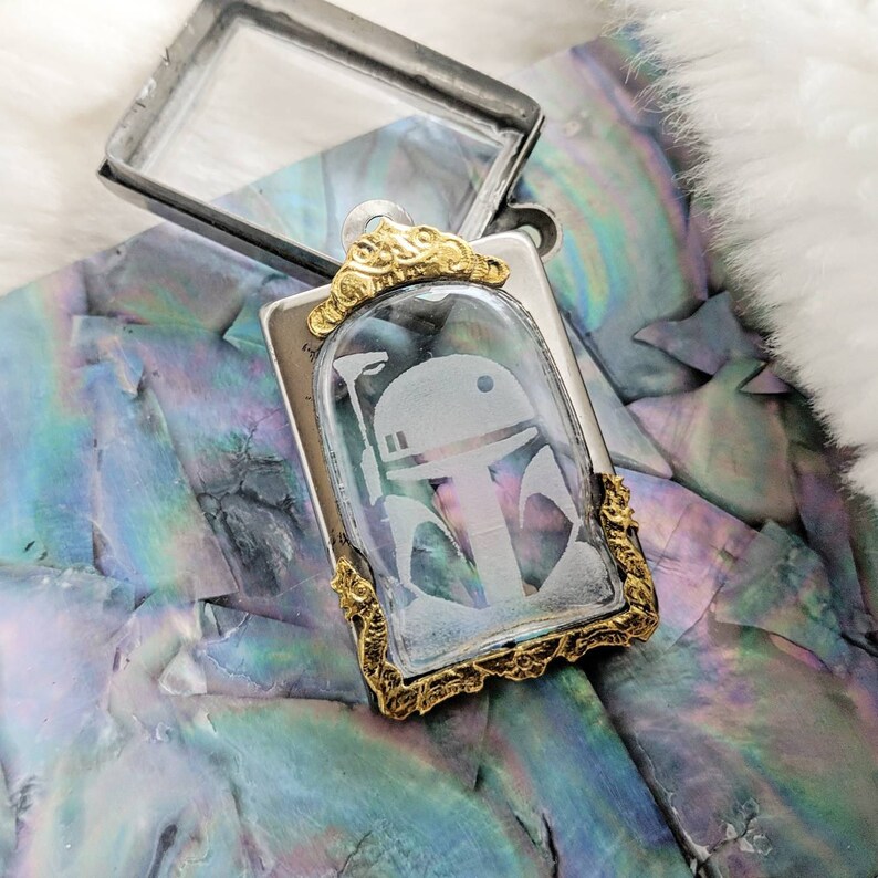 May include: A silver and gold pendant with a clear glass insert featuring an etched image of a Star Wars character's helmet. The pendant is on a white background.
