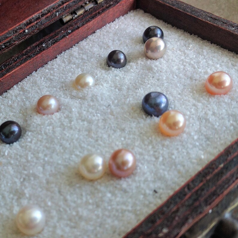 May include: A collection of pearls in various colors, including white, pink, and black, displayed in a wooden box lined with white sand.