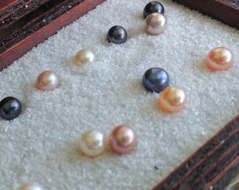 May include: A collection of pearls in various colors, including white, pink, and black, displayed in a wooden box lined with white sand.