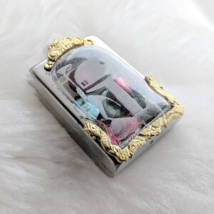 May include: A silver and gold pendant with a clear glass dome containing a small, white, plastic figurine of a Star Wars character. The figurine is surrounded by colorful, iridescent glitter.