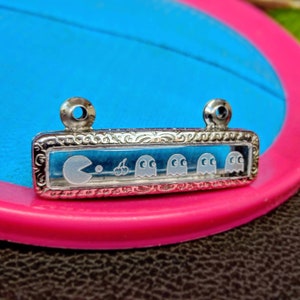 May include: A silver charm with a clear glass insert featuring a Pac-Man character and four ghosts. The charm has a detailed, ornate design around the edges.