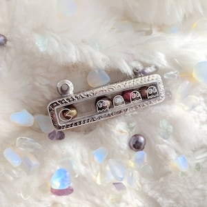 May include: A silver charm bracelet with a clear glass insert featuring a Pac-Man character and four ghosts. The ghosts are in different colors: red, pink, blue, and orange. The charm is on a white fuzzy background.