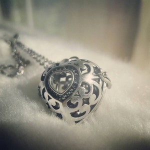 May include: A silver heart-shaped pendant necklace with an intricate openwork design. The pendant features a smaller heart shape in the center, accented with small black stones.