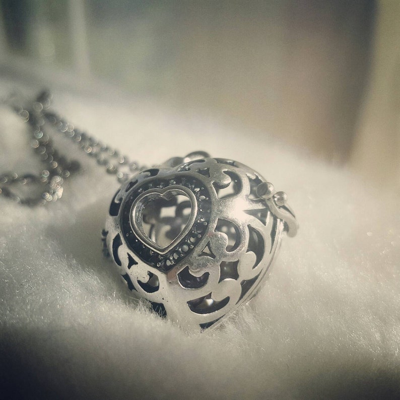 May include: A silver heart-shaped pendant necklace with an intricate openwork design. The pendant features a smaller heart shape in the center, accented with small black stones.