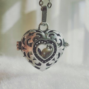 May include: A silver heart-shaped locket necklace with an intricate filigree design. The locket has a small heart-shaped window that reveals two white pearls inside.