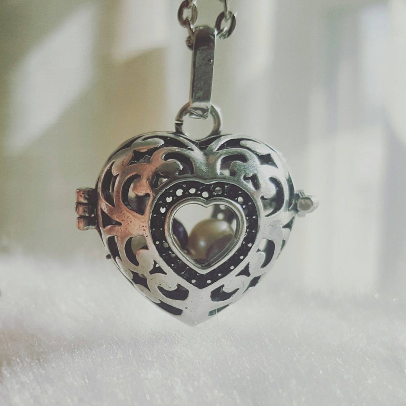 May include: A silver heart-shaped locket necklace with an intricate filigree design. The locket has a small heart-shaped window that reveals two white pearls inside.
