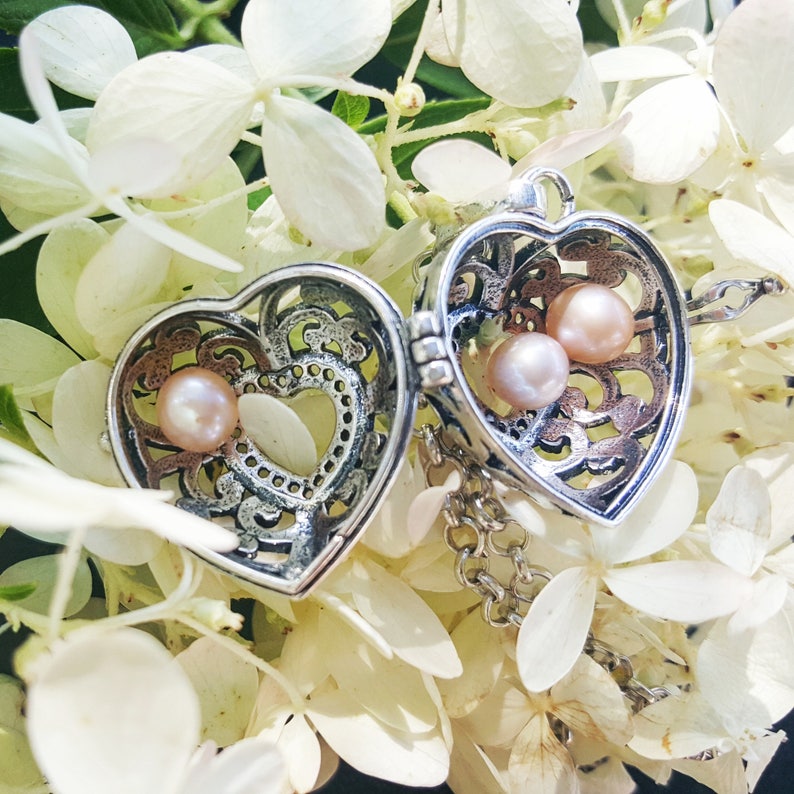 May include: A silver heart-shaped locket with a filigree design and a chain. The locket is open and contains two small pink pearls.