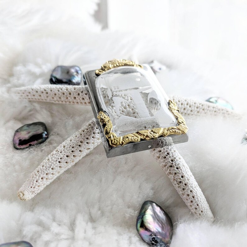 May include: A silver and gold pendant with a clear glass insert featuring a detailed anchor design. The pendant is resting on a white starfish and surrounded by iridescent seashells.