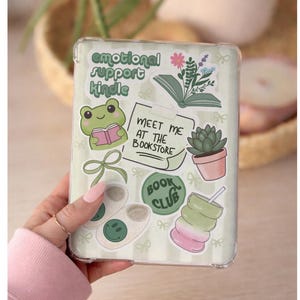 May include: A clear e-reader case decorated with various stickers including a green frog reading a book, a succulent in a pink pot, a green bow, slippers with smiley faces, and text that reads emotional support kindle, meet me at the bookstore, and book club.