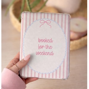 Puede incluir: Una funda transparente para tableta con un diseño de rayas rosas y blancas. La funda presenta un óvalo blanco con un lazo rosa y las palabras "booked for the weekend" en escritura rosa. La funda es sostenida por una persona.