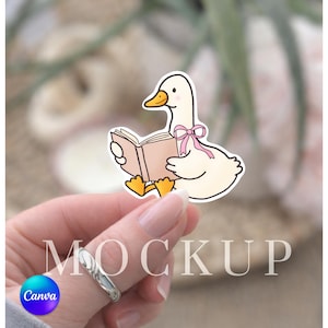 Può includere: Un adesivo con un'anatra dei cartoni animati che legge un libro. L'anatra è bianca con becco e zampe gialle e un fiocco rosa. Il libro è marrone chiaro. La parola "MOCKUP" è stampata nell'immagine.