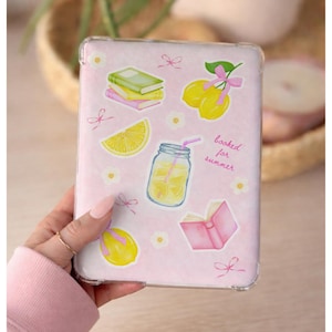 May include: A clear protective case for an e-reader featuring a light pink background with various stickers including a stack of books, yellow lemons with pink bows, a mason jar of lemonade, a pink open book, and small white flowers with text.