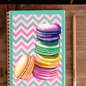 Op de afbeelding: Een stapel kleurrijke macarons op een roze en grijs chevron-patroon notitieboekje. De macarons zijn gestapeld met de grootste aan de onderkant en de kleinste aan de bovenkant. De macarons zijn in tinten van paars, groen, geel en roze.