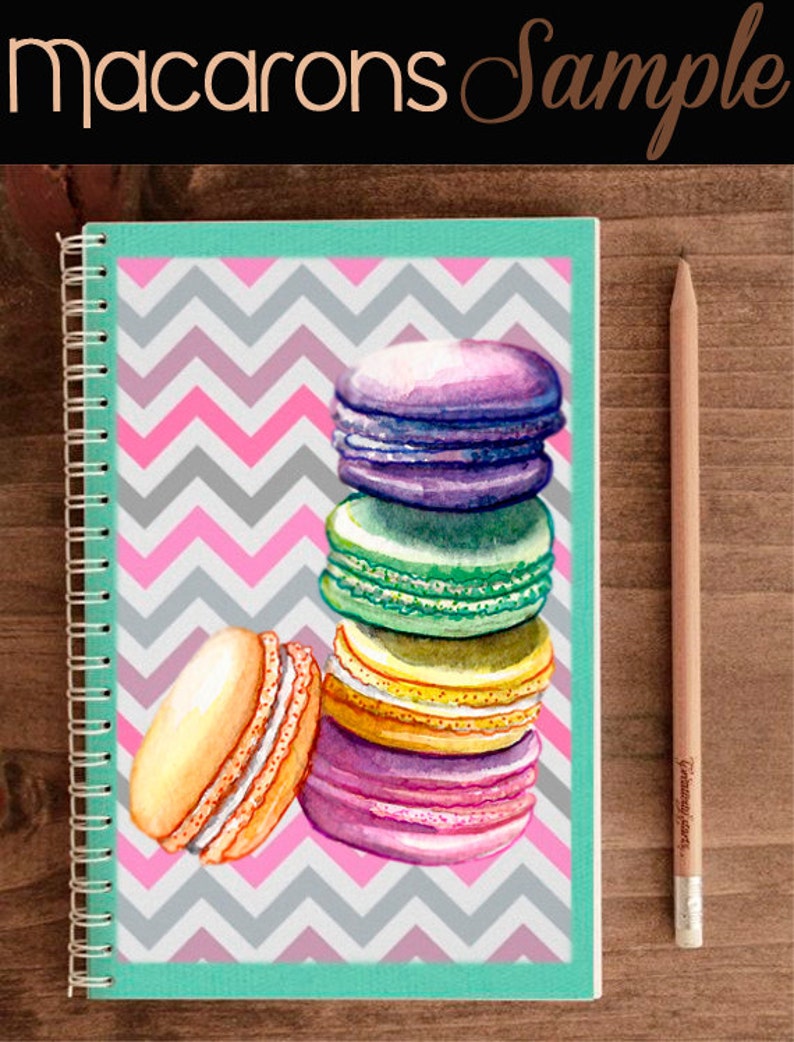 Op de afbeelding: Een stapel kleurrijke macarons op een roze en grijs chevron-patroon notitieboekje. De macarons zijn gestapeld met de grootste aan de onderkant en de kleinste aan de bovenkant. De macarons zijn in tinten van paars, groen, geel en roze.