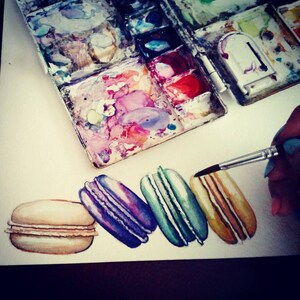 Op de afbeelding: Een aquarel schilderij van vijf macarons. De macarons zijn in een rij van links naar rechts gerangschikt. De macarons zijn geschilderd in tinten beige, paars, groen en geel. De macarons zijn geschilderd op een witte achtergrond.