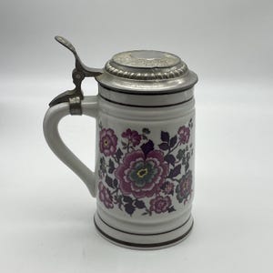 Può includere: Un boccale da birra in ceramica bianca con un motivo floreale nei toni del viola, rosa e verde scuro. Il recipiente presenta un coperchio metallico incernierato con leva e un manico curvo, arricchito da sottili bande orizzontali scure.
