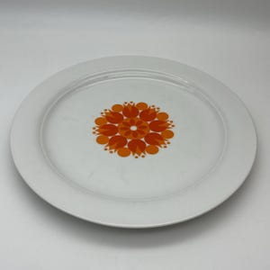 Op de afbeelding: Witte keramische plaat met een levendig oranje bloemmotief in het midden. De plaat heeft een brede rand en een glad oppervlak. Het bloemmotief is symmetrisch en heeft gestileerde bloemvormen.
